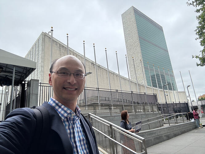 Man in suit smilng for selfie in fron of a wide building with flags in front plus a towering glass building on the far right