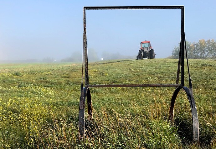 An open rectangular metal frame mounted on metal wheels sits in an open field, through the frame is a tractor driving towards the horizon