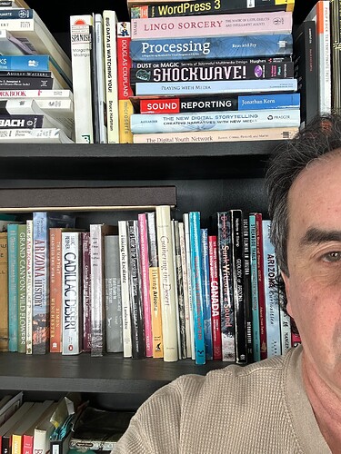 A dark haired main standing half off screen on the right (we see only one eye, hair, and. a shoulder) stangind in front of two shelves of books randomly stacked. the top shelf includes WordPress 3, Shockwave! and more technology books. The second shelf includes Arizona History, Cadillac Desert, an Edward Abbey book, other titles.