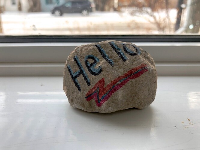 A round rock with handwritten large letter Hello sits on a window sill with a snowy street scene seen outside.
