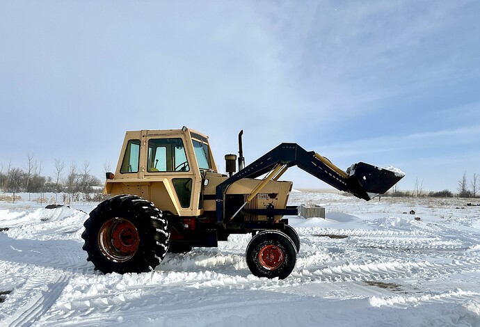 This image depicts a tractor operating in a snowy field. The tractor is equipped with a front loader attachment, which is currently raised and appears to be holding snow. The tractor has large tires, suitable for navigating through snowy terrain. The background shows a flat, open field covered in snow, with some trees and a clear sky visible in the distance. The scene suggests that the tractor is being used for snow removal or other winter-related tasks.