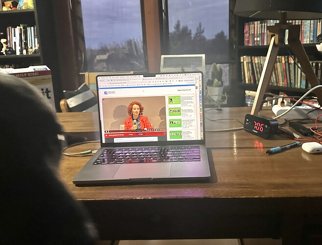 Laptop computer on desk with live stream keynote, on the left is blurred face of a black dog. A clock on right reads 7:06am