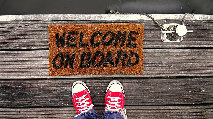 View of two feet in red shoes with white laces standing on a wood boat deck. At top is a floor mat reading Welcome on Board