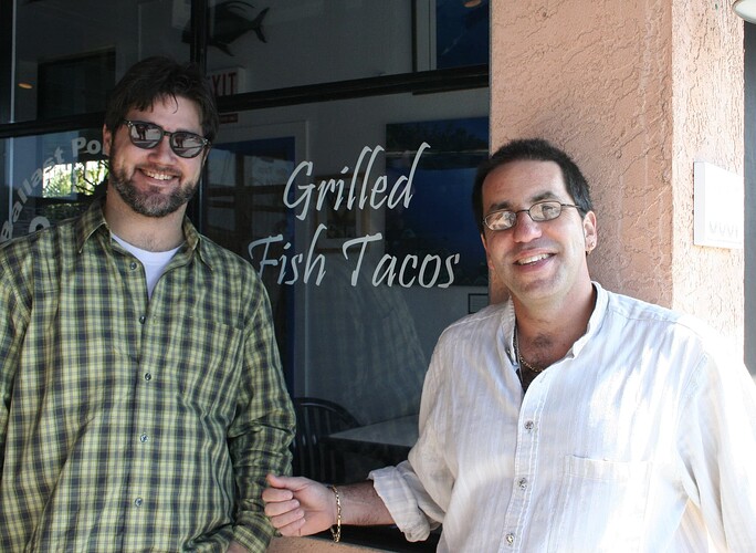Two men smiling outdoors in front of a restaurant window advertising "Grilled Fish Tacos".