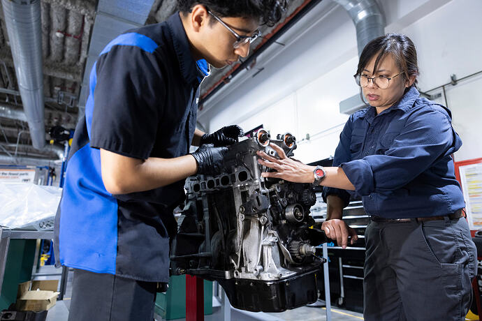 Asian female instructor working on an engine with a Hispanic male student in auto shop on a college campus.