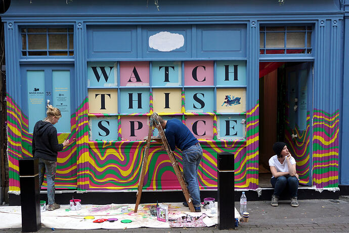Woman and man painting multiple curveing lines of color on a building front where large letters in squares read Watch This Space