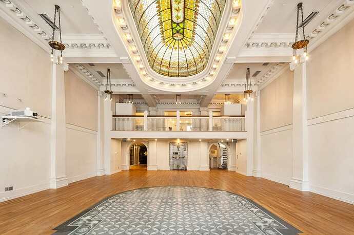 Interior view of a spacious room with a stained glass dome ceiling, polished wooden floor, and decorative railing. A patterned mosaic is visible on the floor, and there is a staircase on the right.l
