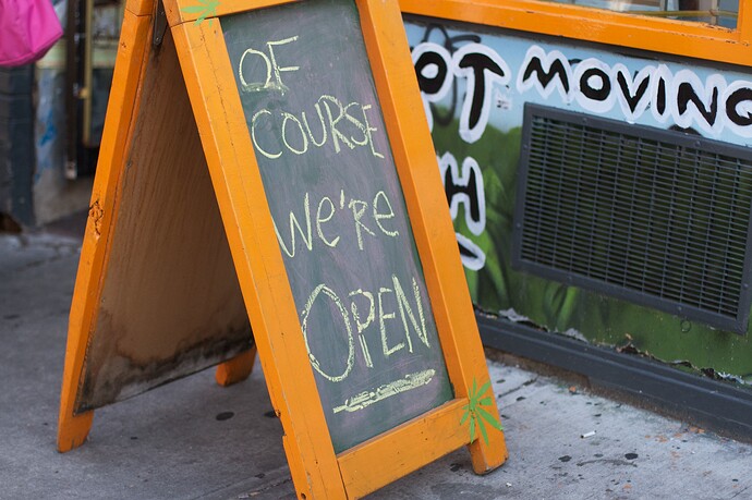 Orange A-frame chalkboard sign reading “OF COURSE WE’RE OPEN” outside a storefront.