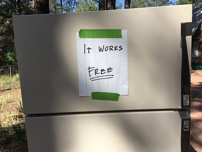 This image shows a refrigerator placed outdoors. Attached to the front of the refrigerator is a handwritten note on a piece of lined paper. The note reads, "It works. Free." The note is secured to the refrigerator with green tape. The background shows a natural setting with trees and grass, indicating that the refrigerator is likely being offered for free to anyone who wants it.