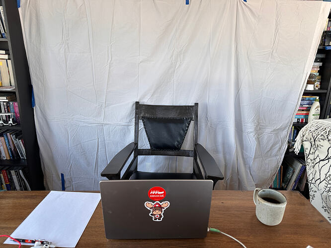 Back of an open laptop atop a wood desk, behind is an empty old wood chair with large armrests. A white sheet is hanging behind the chair covering the books on shelves.