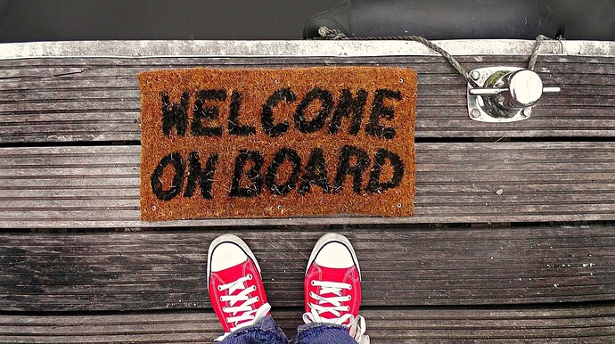 A doormat on a wooden deck with "WELCOME ON BOARD" in black letters. Red sneakers are visible at the bottom of the image.