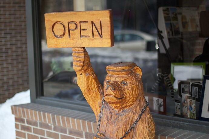 Small wooden bear statue in front of a building window with hand is raised, holding a sign that reads Open