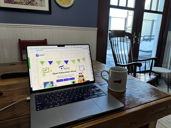 Laptop on wood table open to the Open Education Week web site. A coffee cup sits next to the laptop. Beyond to the right is a wooden rocking chair in front of glass french doors through which you can see snow piles two feet high.