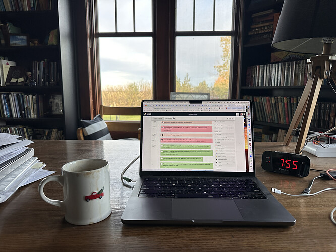 Open laptop with conference schedule on screen, sitting on a wood desk with a coffee cup on the left, a clock reading 7:55am on right, stray cables and piled papers to the side, behind is a window with trees showing fall colors