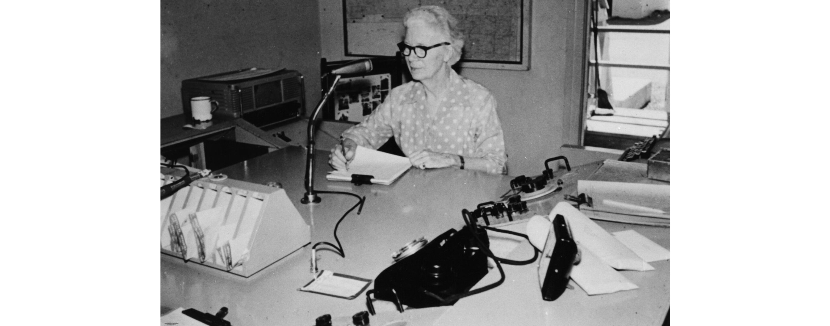 Old photograph of woman sitting at large desk, reading from notes, speaking into a microphone with radio equipment behind her