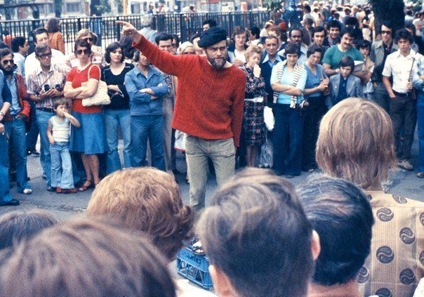 Man in beret points emphatically while a crowd listens behind and in front of him, the clothing is definitely from the 1970s