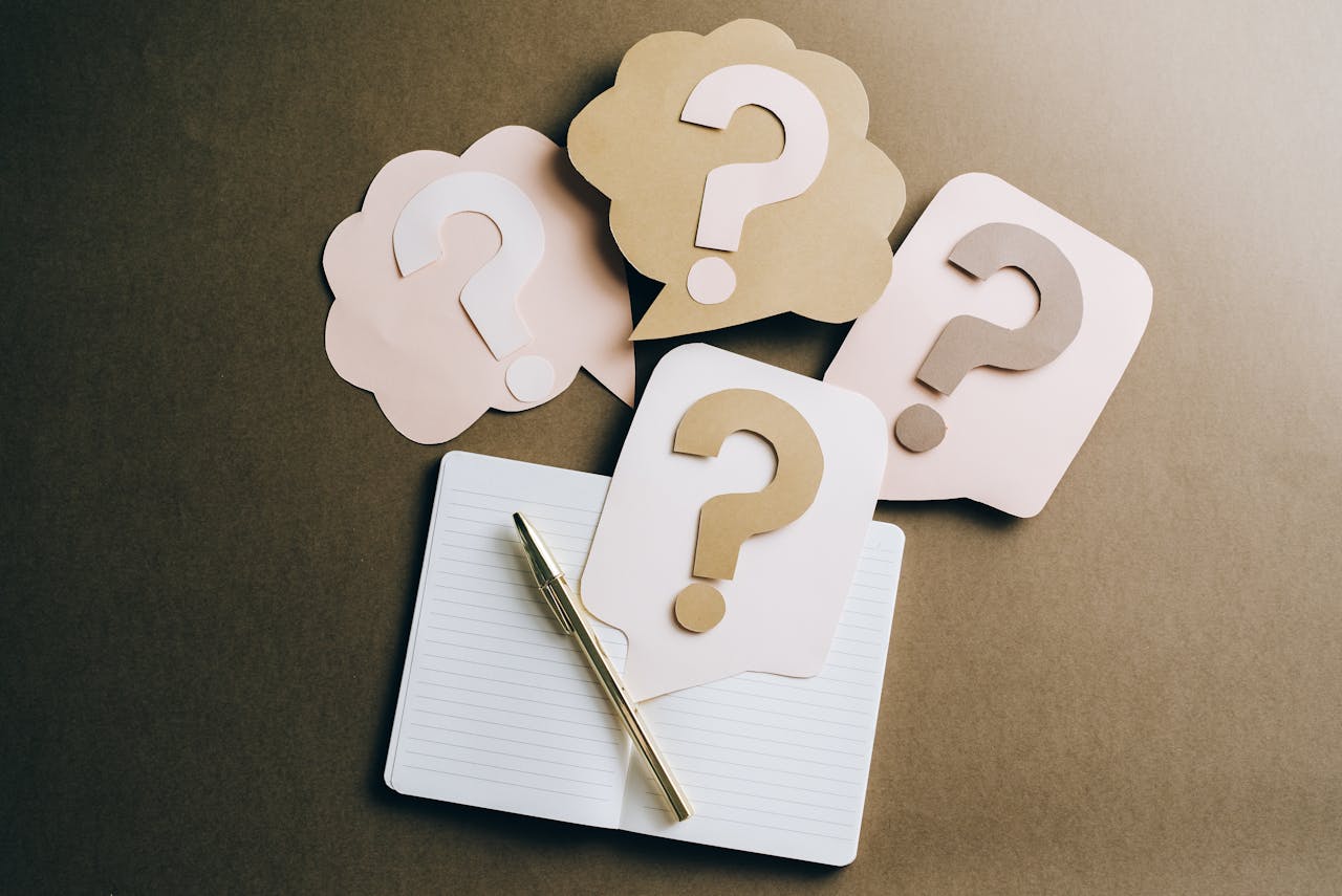 Four Large question marks each cut out of paper embossed on shapes of thought bubbles, spread on a table over an open blank notebook with a pen.