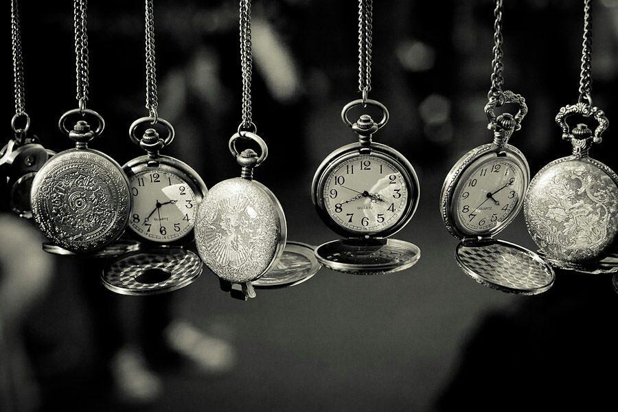 A row of seven old silver watches hanging on chains, 3 of them with clocks facing, others with their backs of detailed pattern engravings, all in front of a blurred dark background perhaps in a shop