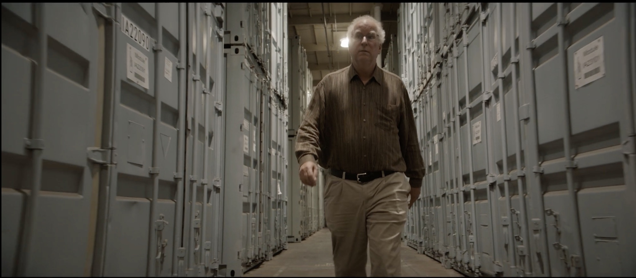 A man walking through a dimly-lit storage space. The corridor is flanked by large, light blue metal containers, each secured with latches..